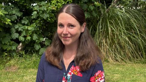 A young woman with long dark hair over both her shoulders wearing a dark blue top with flowers on it, standing in front of a hedge in a garden.