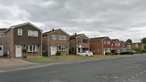 A suburban residential street lined with detached houses on one side under a cloudy, overcast sky. The viewpoint is taken from across a wide road, looking diagonally down the street as it stretches into the distance on the right-hand side.