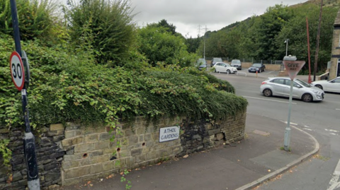A brick wall in a state of disrepair. A street sign reads Athol Gardens. Beyond is a busy main road