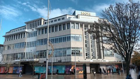 A large building in Hull city centre. It has lots of windows and is opposite Hull train station. The architecture is impressive and Hammonds of Hull is written on multiple signs, however the bottom floor os boarded up and hasn't been occupied for three years. 