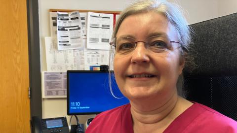 Dr Helen Jervis, who has grey hair, glasses and is wearing fuchsia-coloured scrubs, sitting in front of her monitor and telephone in the Temple Sowerby GP surgery.