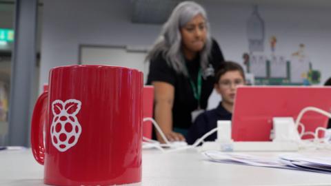 A red Raspberry Pi-branded mug sits in the foreground of a tech-focused workspace. Behind it, two (an adult and a child) are engaged with a computer screen. The adult has long grey hair and is helping the child with their work.