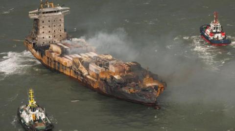 Smoke emitting from a semi-burnt out cargo vessel, which is drifting in the sea. Two smaller vessels are on either side of it 