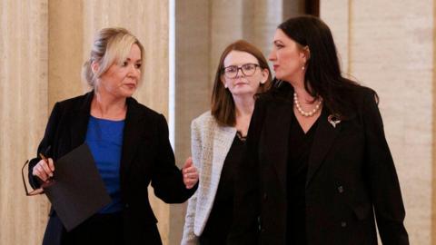 From left to right of the image, Michelle O'Neill, Caoimhe Archibald and Emma Little-Pengelly stand in front of mics inside Stormont