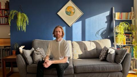 Male sitting on sofa wearing T shirt and dark trousers