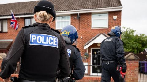 Three police officers taken from behind in riot gear one holding a battering ram as they arrive at a house to execute a warrant