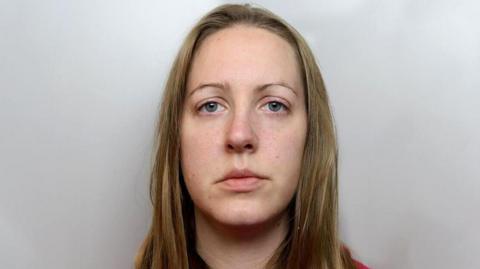 Lucy Letby police mugshot. Sandy blonde hair is worn long over a red hoody. She has thin eyebrows and bluey grey eyes.