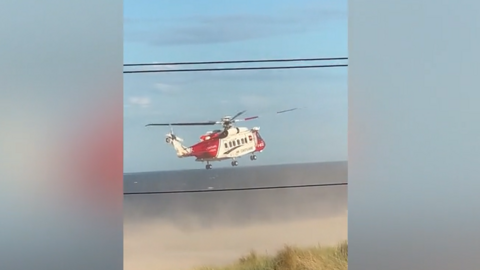 A red and white helicopter trying to land on the beach. Sand has been whirled up around the craft and into the sky by the downwind from the blades.