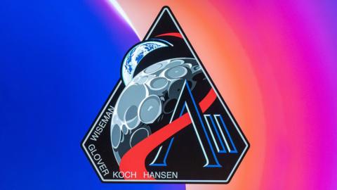The emblem is the shape of an upside down diamond. Its background colour is black and has a grey line around the edge. In the bottom left hand corner of the patch are the names Wiseman, Glover, Koch and Hansen. In the middle of the patch is a cartoon moon. There is a red wave line going over it. It also goes through a black and dark blue AII. Behind the moon and sticking out of the diamond shape is a cartoon image of the Earth