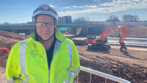 Terry Robinson in a high vis jacket and white helmet. He is smiling at the camera, and there is a digger and the bridge in the background. 