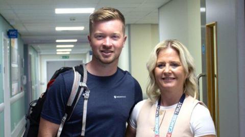 A young man with fair hair and a blue t-shirt and a woman with blonde hair and a beige vest over a white t-shirt stand in a hospital corridor. They are both smiling and standing close together.

