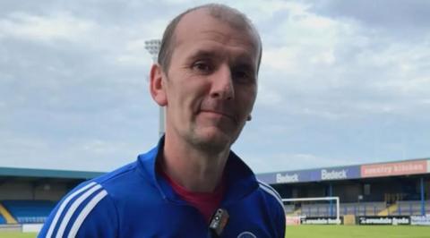 A man looks into the camera. He is wearing a blue tracksuit top with the Glenavon badge and white stripes on the sleeves, and a red t shirt. He has balding brown hair. A small football stand with blue seats and a football pitch is in the background.