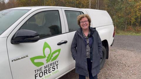 Professor Eunice Simmons has short brown hair and is wearing dark rimmed glasses and a blue coat and is stood next to a white van with The Mersey Forest logo on.