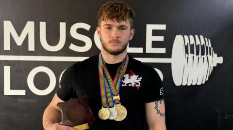 Lucas Williams in a black Cymru Wales team T-shirt, wearing two gold and a silver medal and holding a wooden Commonwealth championship shield. He has a beard and a tattoo on his right bicep showing and is standing in front of a gym logo showing a barbell image