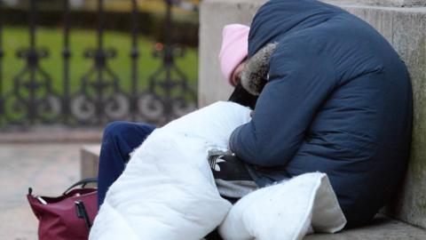 Two anonymous people huddled on a concrete step in what appears to be a park. One is wearing a pink hat and has a pink bag. The other is wearing a blue coat and is covered with a blanket.