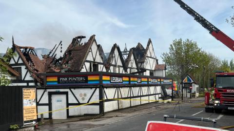A nightclub, pictured in the middle of the day, with a roof that has been destroyed in a fire. A fire engine is parked opposite the venue. There is a long aerial ladder rising into the sky above the building. Some smoke can be seen billowing out of the venue.