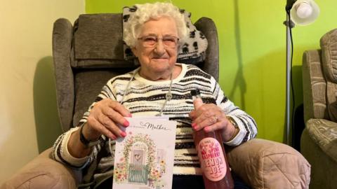 Joyce Lynas sits in a grey armchair, smiling at the camera. She has short, white hair worn in a perm, and wears a white jumper with black stripes and a long silver necklace. She holds a birthday card and a bottle of pink lemonade.