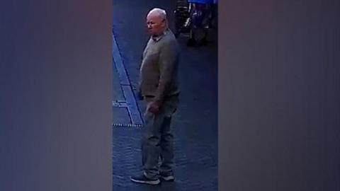 A blurred CCTV image of a white man with balding grey hair. He is wearing a jumper, shirt, chino trousers and blue shoes. He is standing on a pavement.