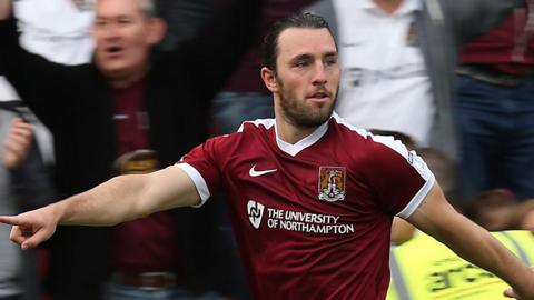 John-Joe O'Toole of Northampton Town celebrates after scoring his sides second goal. He is pointing with one hand and is looking to the sign. Out of focus fans behind are celebrating.
