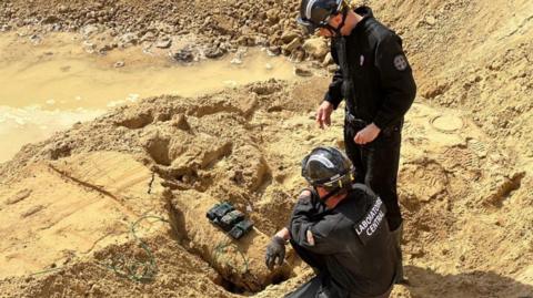 Two bomb disposal experts stand over a WW2 bomb that has been rigged with explosives, surrounded by sand.