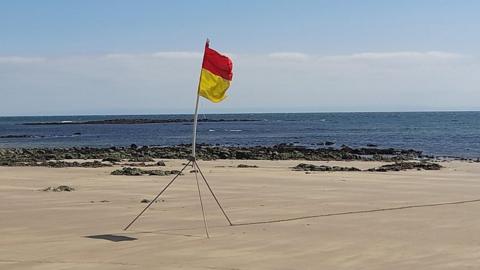 A red and yellow flag sits on the sand at the Mumbles, Wales, with blue sea behind and a blue sky with some white cloud above