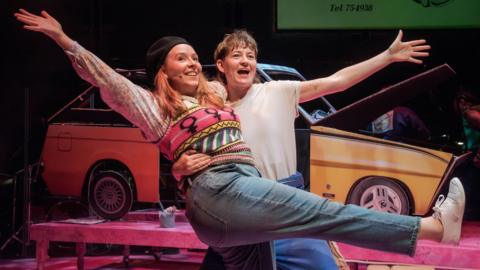 Two performers on stage in a playful pose, with one lifting the other who has arms outstretched and one leg raised. Behind them is a yellow car with its hood open, along with stage props including a bench and an old-fashioned telephone, suggesting a theatrical performance with vibrant lighting.