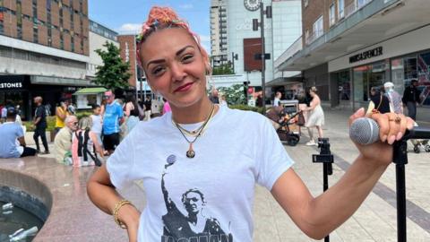 A woman with pink hair is wearing a white t-shirt and is holding a microphone. She is standing in a street full of people walking about and sitting nearby.