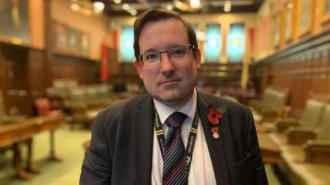 Lawrie Hooper, who is wearing glasses and a dark suit and tie. He is standing in the House of Keys Chamber and is wearing a red poppy.