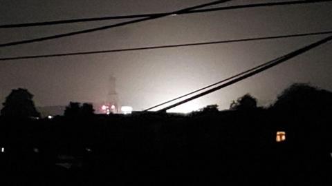 Lights illuminate the sky. In the foreground are silhouettes of trees and electricity wires.