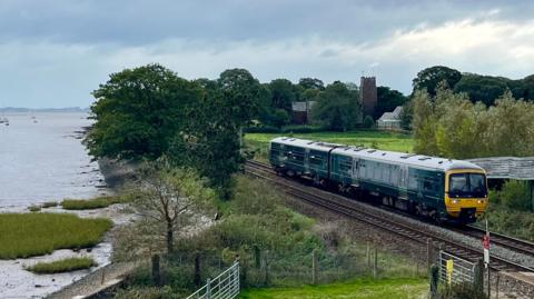 A train heading north on tracks along the Exe Estuary at Powderham with St Clement's Church in the background.