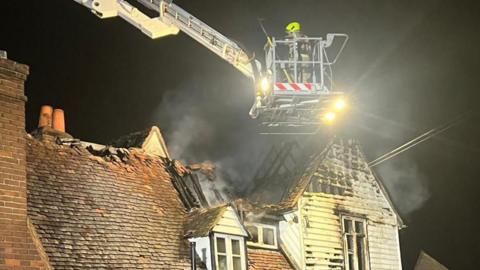 A firefighter is standing on the platform of an aerial ladder above the pub. Smoke is coming from the roof, part of which is charred and has collapsed. It is dark and the sky is black