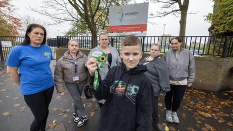 A picture of student Finlay holding up his sunflower lanyard in protest with some parents outside Werneth School on Harrytown road in Stockport