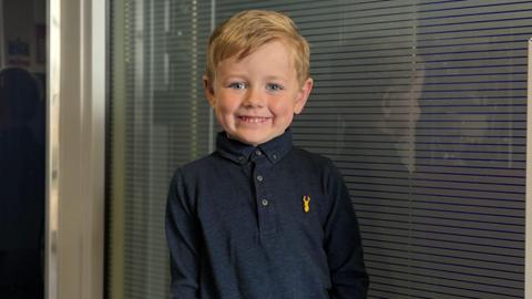 A young boy with dark blonde hair is smiling at the camera. He is wearing a dark navy shirt with a yellow logo on the breast in the shape of a stag's head