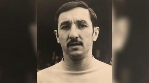 A black and white headshot of Rod Thornhill. He is wearing a light-coloured top and has dark hair and a dark moustache. He's looking at the camera and frowning slightly.