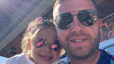 A man wearing sunglasses smiling with his young daughter beside him