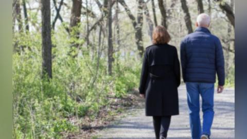 A man and a woman are walking on a path through woodland. The woman had auburn hair and a black knee-length coat and the man has grey hair, a blue padded jacket and jeans. They are walking away from the camera.