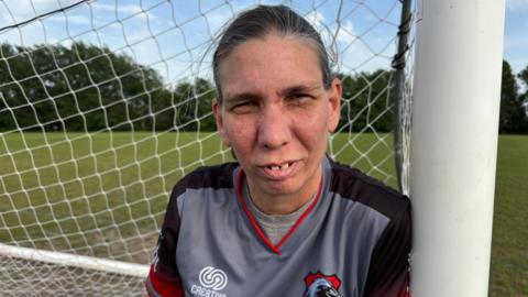 A lady in her 40s leant against a goal post with frowned eyes and a stern face wearing a Ravenshead Reds football shirt.
