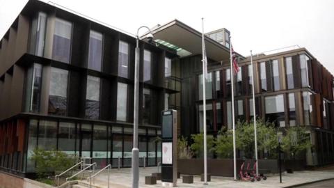 An office building with glass doors, and two rows of vertical windows above the doors. Three flag poles stand in the square at the front of the building. One flies a council flag, the middle flies a Union Jack, and the third is empty. Three red scooters stand on the pavement next to the flag poles. 