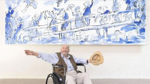 Quentin Blake, sat in a wheelchair, wearing a light blue shirt and brown waist jacket and holding a tan hat with his arms outstretched under a large blue and white mural.