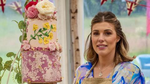 Georgie Grasso stood in the Bake Off tent. She is wearing a beige apron and underneath it has a blue, pink and white marbled top. She is looking at the camera with her brown and blonde hair half tied up and half down. There is a cake to the left of her, which is a three tiered cake, in different shades of pink with flowers adorned on it.