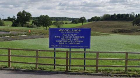 A blue sign saying Woodlands Golf and Country Club, above a fence with green grass and trees behind.