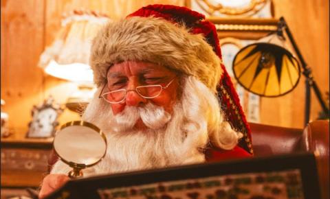 Santa who has a traditional red and white hat, white beard with half rimmed glasses is holding a magnifying glass over his Christmas list.