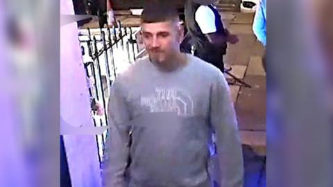 CCTV image of a man in a bar. He has dark hair and is wearing a grey North Face jumper with dark blue jean