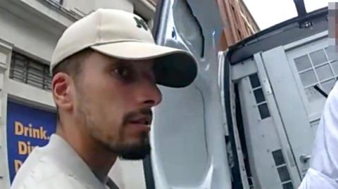 Brahim Kaddour-Cherif seen in arrest footage from police bodycam. He is wearing a baseball cap and white sleeveless coat with a grey top on underneath. He is being led into a police van by an officer wearing body armour.