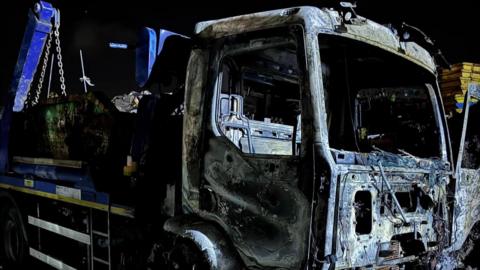 A burned-out lorry at the Kendrick Industrial Estate. It is dark and the entire front of the vehicle has been burned, with missing windscreen 