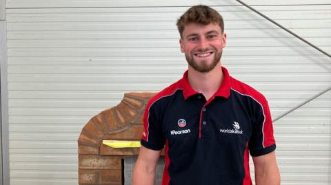 Joe Shingler, wearing a WorldSkills black and red polo shirt. He has short ginger hair and beard and is smiling at the camera against a white backdrop, with an example of his brickwork behind him. 