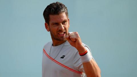 Cameron Norrie is wearing a white t-shirt with a red sash and holding up his fist