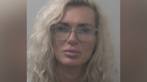 Kasha Sanderson has wavy blonde hair and is wearing glasses and makeup in a police mugshot.