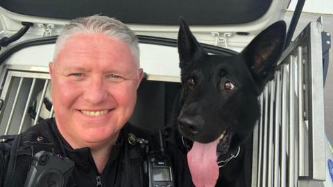 A police officer smiling at the camera with a black German shepherd, who is sticking his tongue out