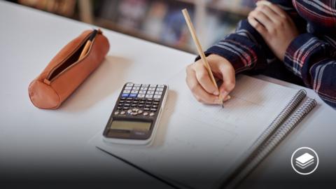 A photograph of a students hand writing on maths paper with a pencil, with a pencil case and calculator next to them.